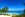 A sunny day at Grace Bay Beach in Providenciales, Turks and Cacos. A walkway on a white sandy is bordered by palm trees and plants. The calm ocean stretches out towards the horizon under a deep blue sky.