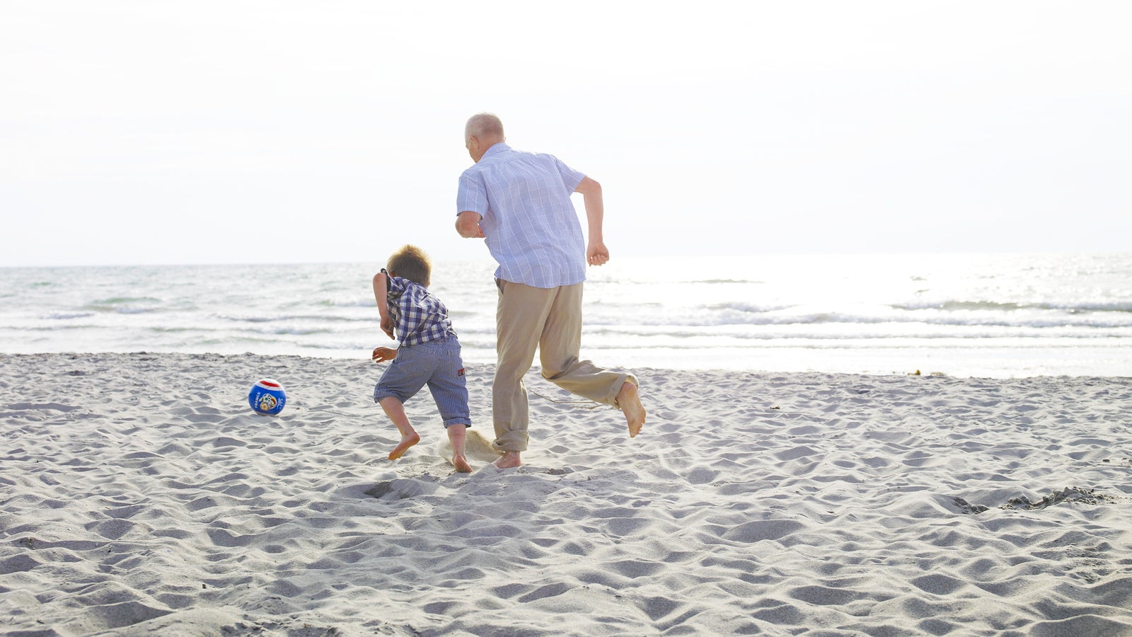 Grandfather playing football on beach with grandson (4-6) rear view