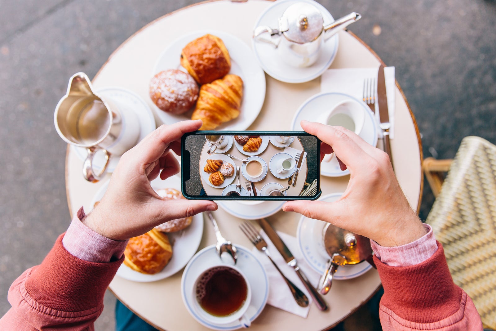 Young man photographing French breakfast with croissants on the table in sidewalk cafe with smartphone, Paris, France