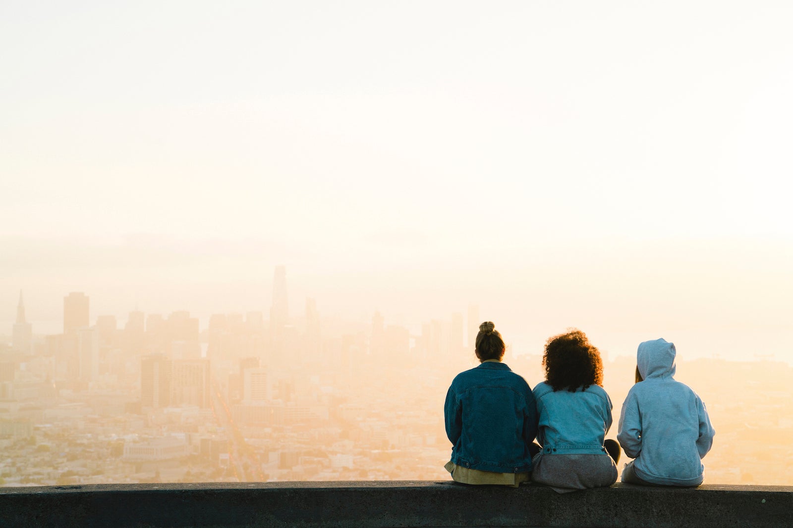 Rear view of female friends looking at cityscape while sitting against sky during sunset