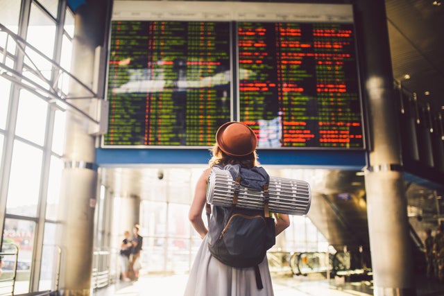 Theme travel public transport. young woman standing with back in dress and hat behind backpack and camping equipment for sleeping, insulating mat looks schedule on scoreboard airport station