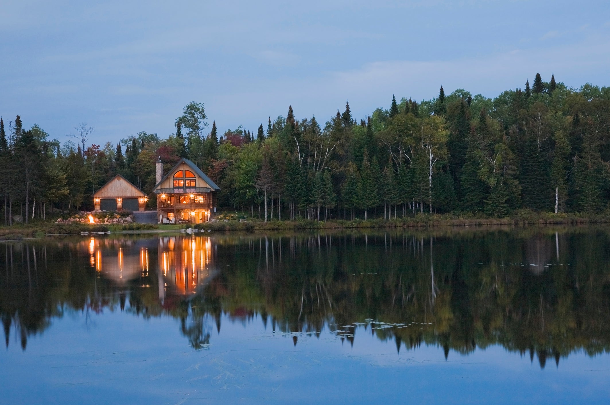 Illuminated garage and handcrafted spruce log home on edge of lake at dusk, Quebec, Canada
