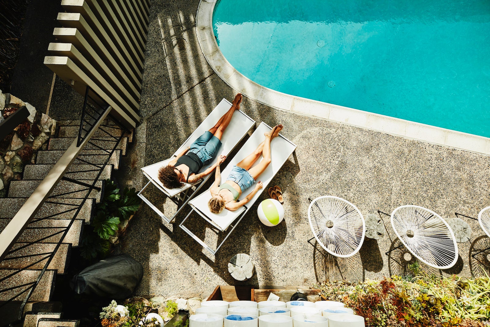 Overhead view of lesbian couple holding hands while relaxing in lounge chairs by hotel pool