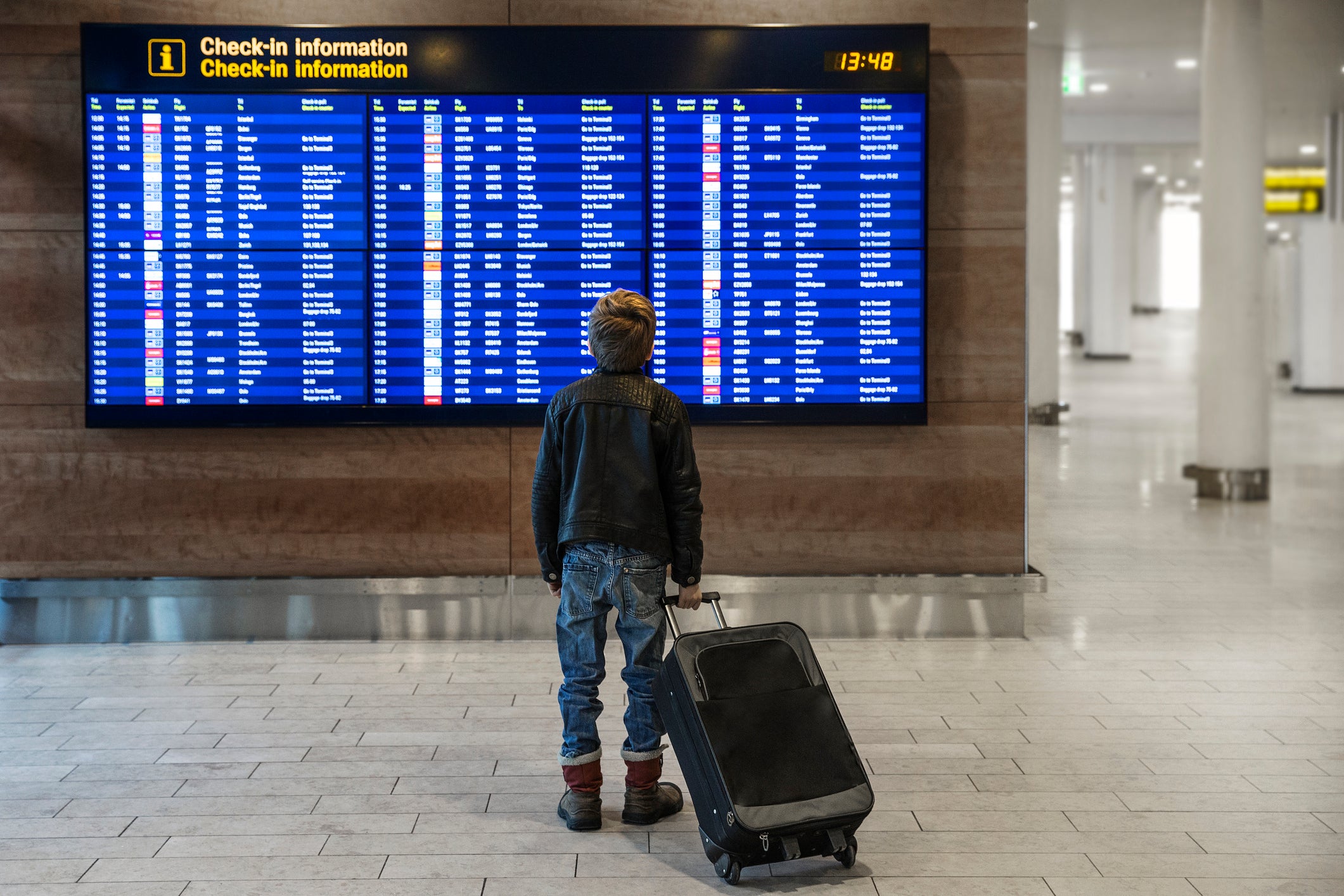 Rear view of boy with wheeled suitcase looking at airport departure board