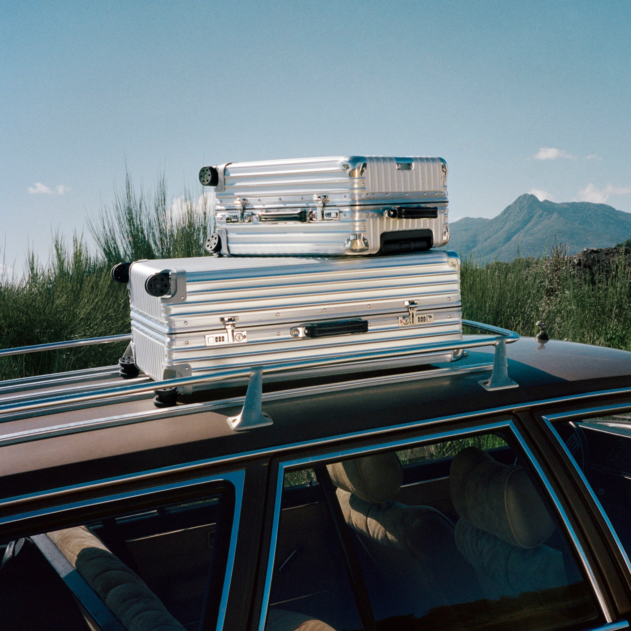 Two silver Rimowa suitcases strapped to the top of a car.
