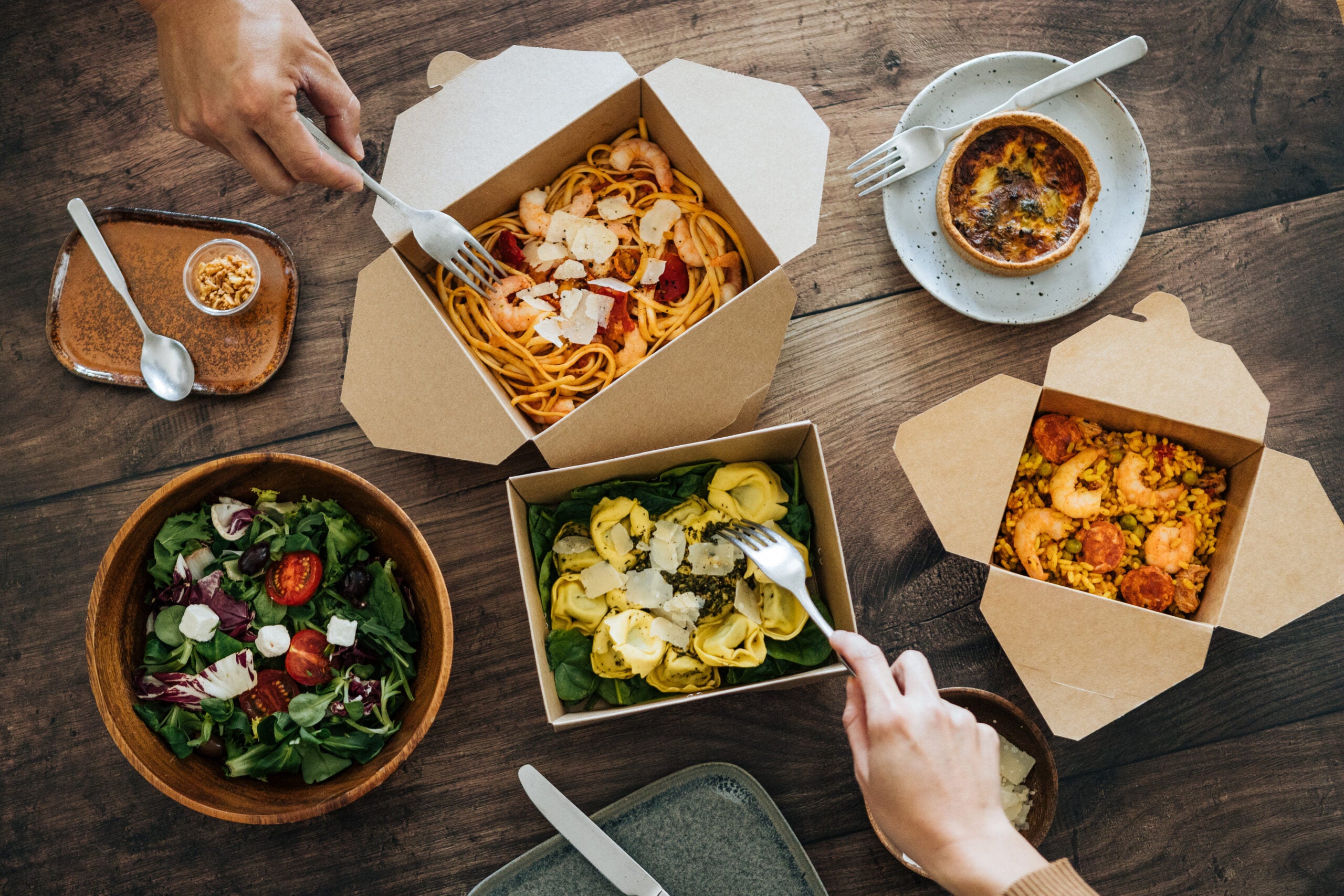 Meals in takeout boxes are open on a table while two unseen people reach for them with forks