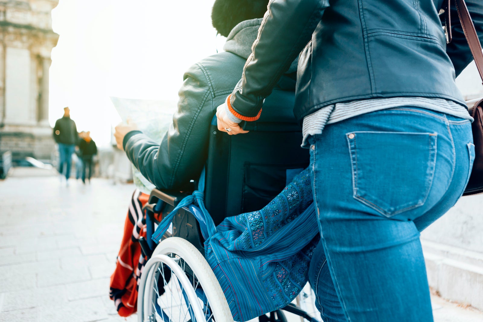Side View Of A Man Sitting In A Wheelchair While A Female Person  Is Pushing Him Around The City