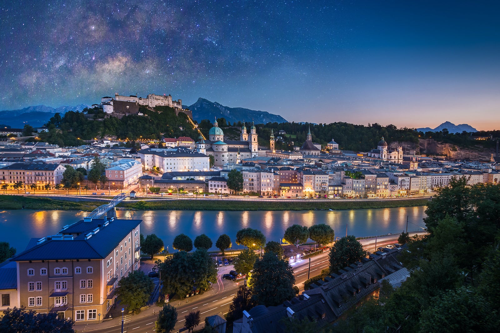 View of cityscape of Salzburg Cathedral, Fortress Hohensalzburg, and old castle in center of old town with river and road along the river at sunset time with milky way in Salzburg, Austria, Europe and also view of snow on alps mountain in background