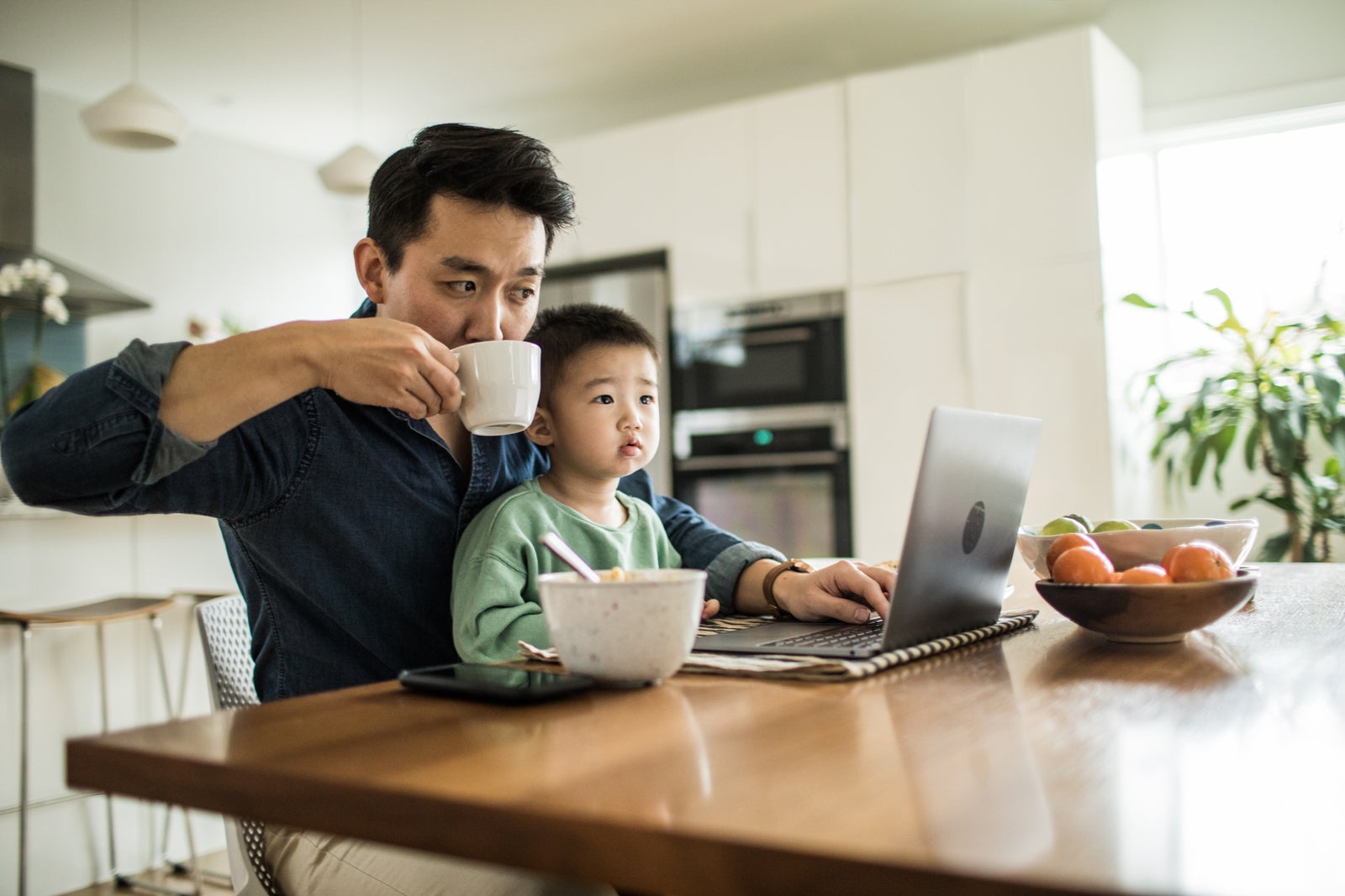 Father multi-tasking with young son (2 yrs) at kitchen table