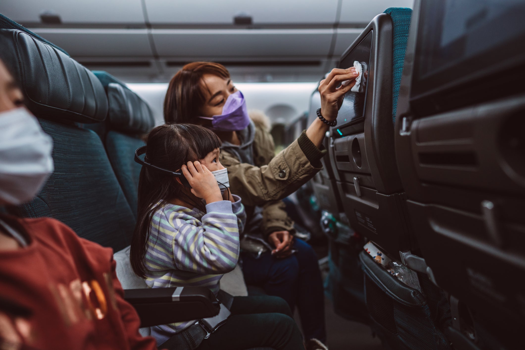 Mom &amp; daughter in protective face masks cleaning the seat-back TV screen with disinfectant surface wipe while travelling on the airplane