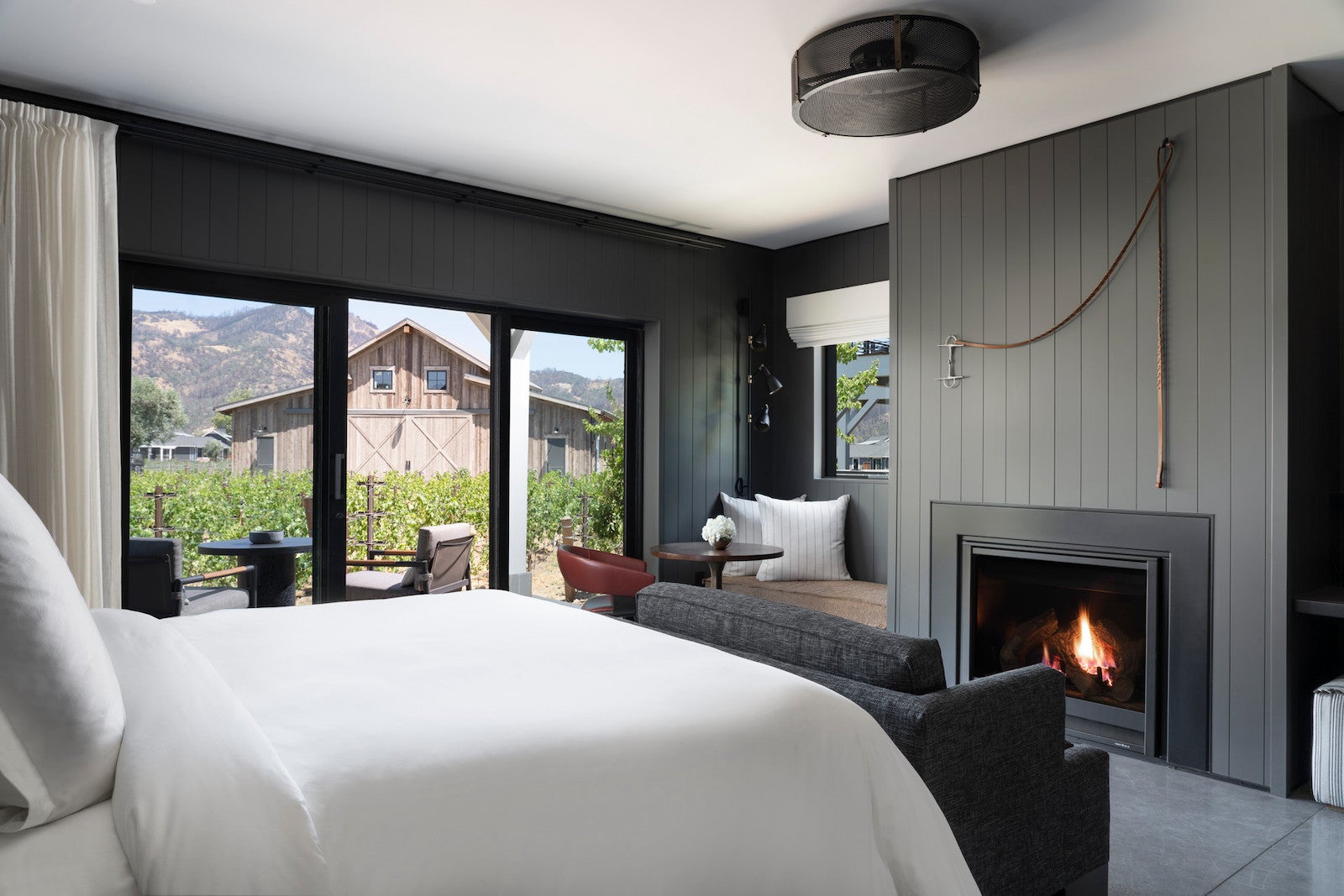 large hotel room with dark wood fireplace, large white bed, and windows looking out onto vineyard and wooden barn
