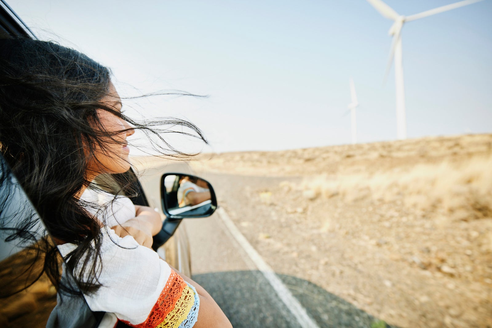 Smiling teenage girl with head out car window and hair blowing in wind passing wind turbines in desert
