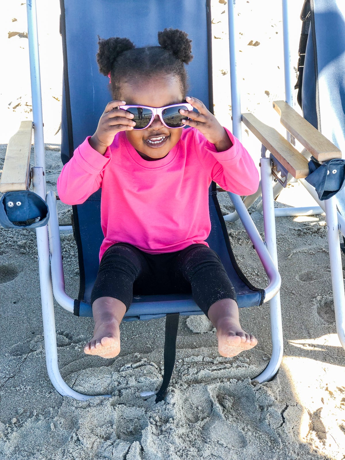 Toddler wearing a long sleeve pink shirt and black leggings sits in a beach chair with pink sunglasses.