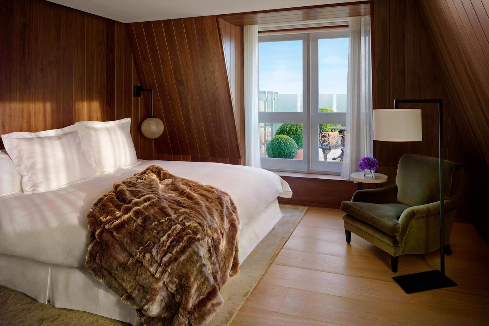 hotel room with white bed, fur blanket, wooden paneled walls and window looking out at trees