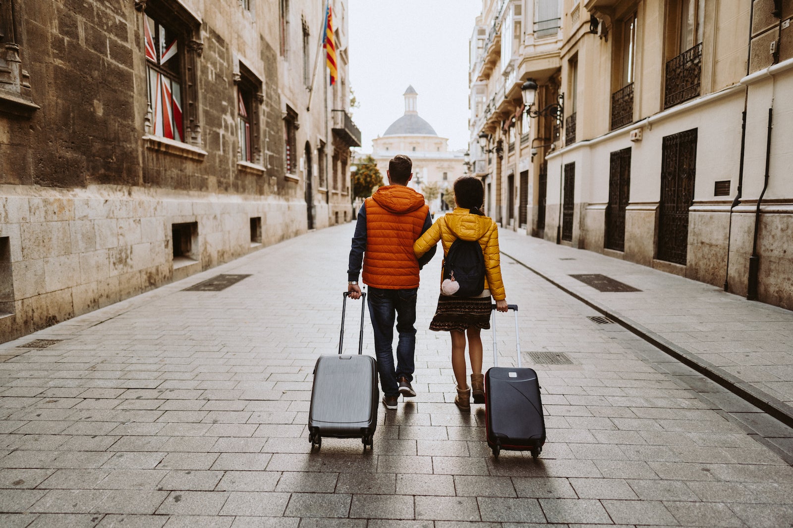 A view from behind of a couple walking on cobble streets with two carry-on sized luggage with wheels.