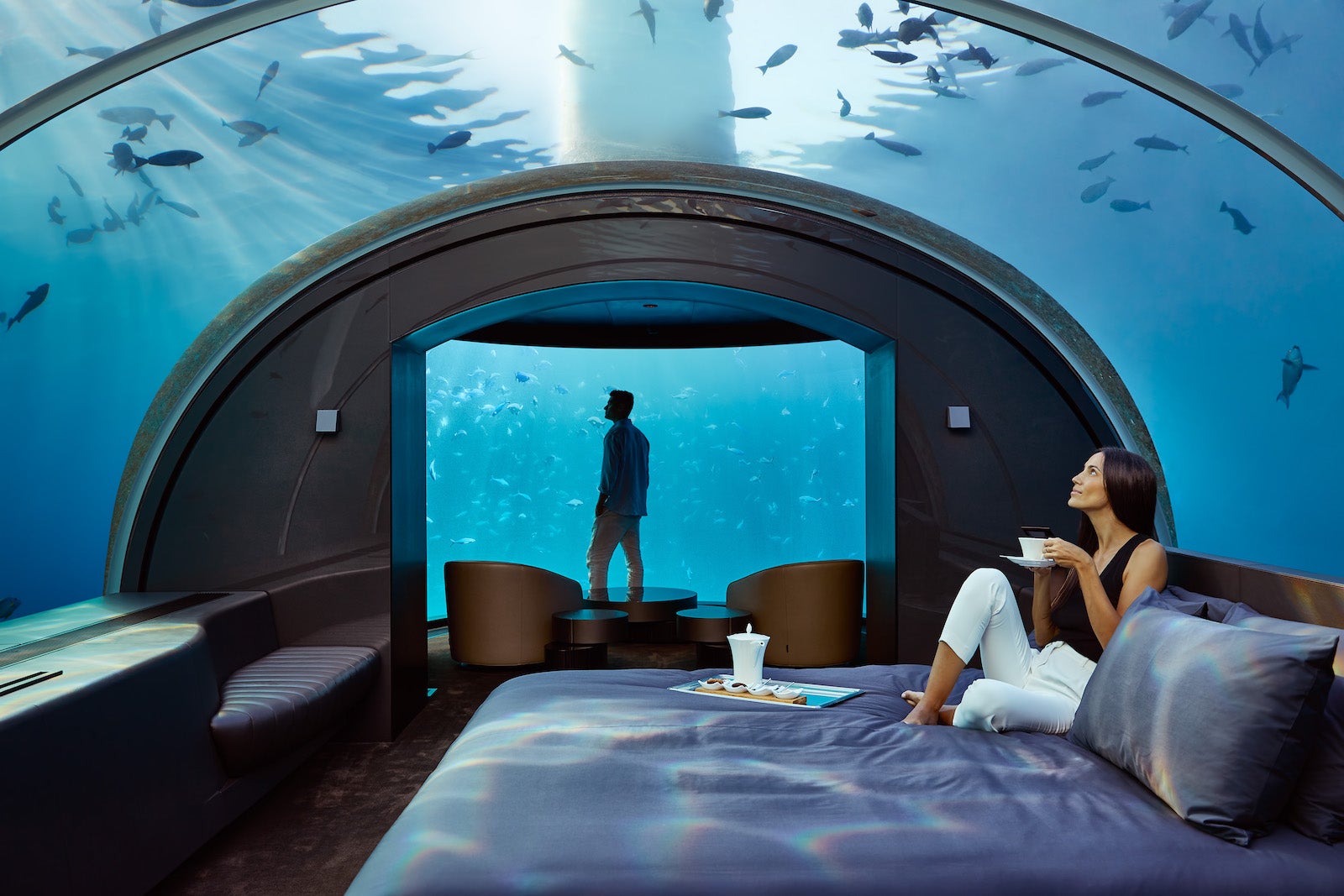 woman sitting on bed in underwater hotel room surrounded by fish