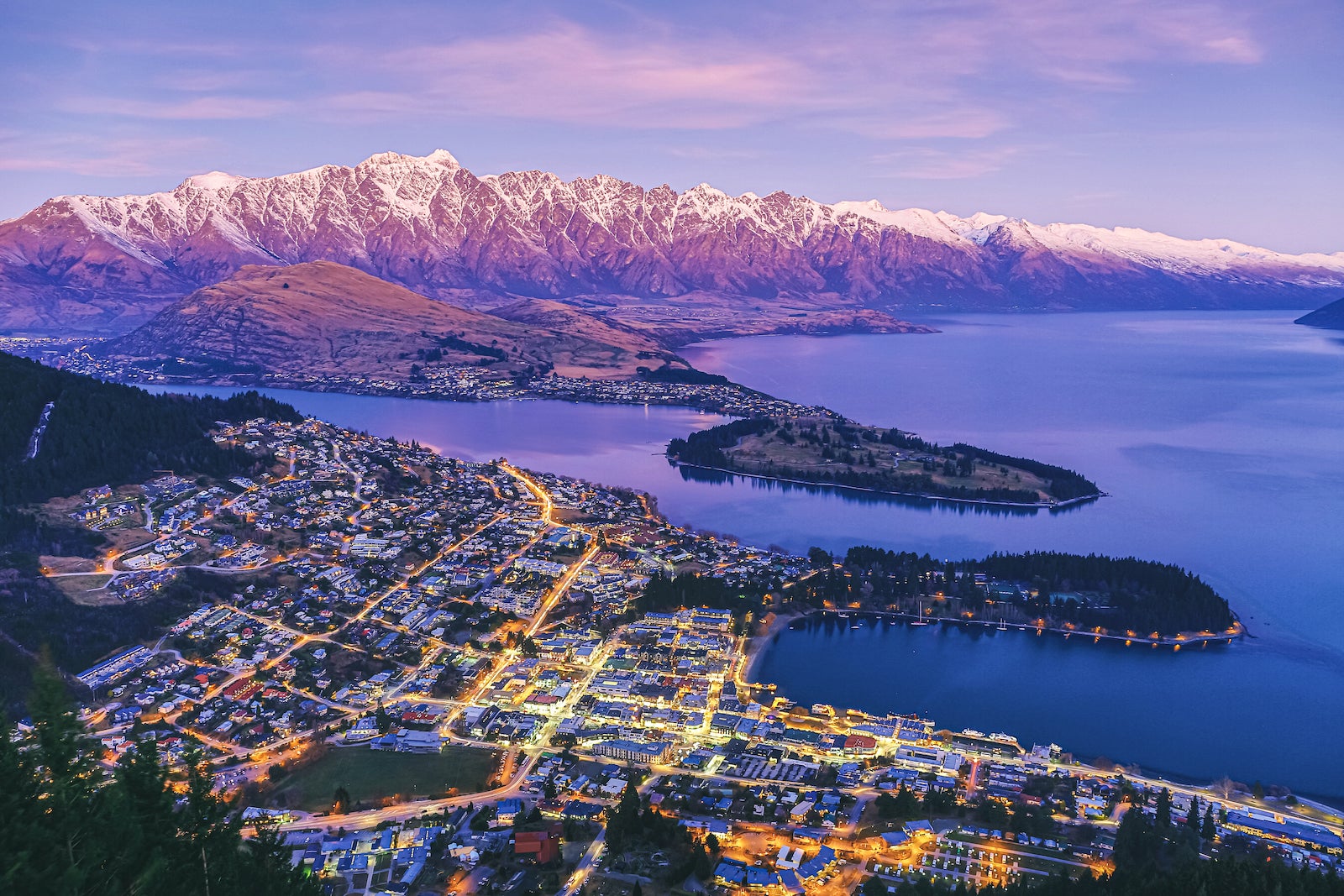 aerial photo of Queenstown Zealand at night with glowing streets and mountains at sunset in the back