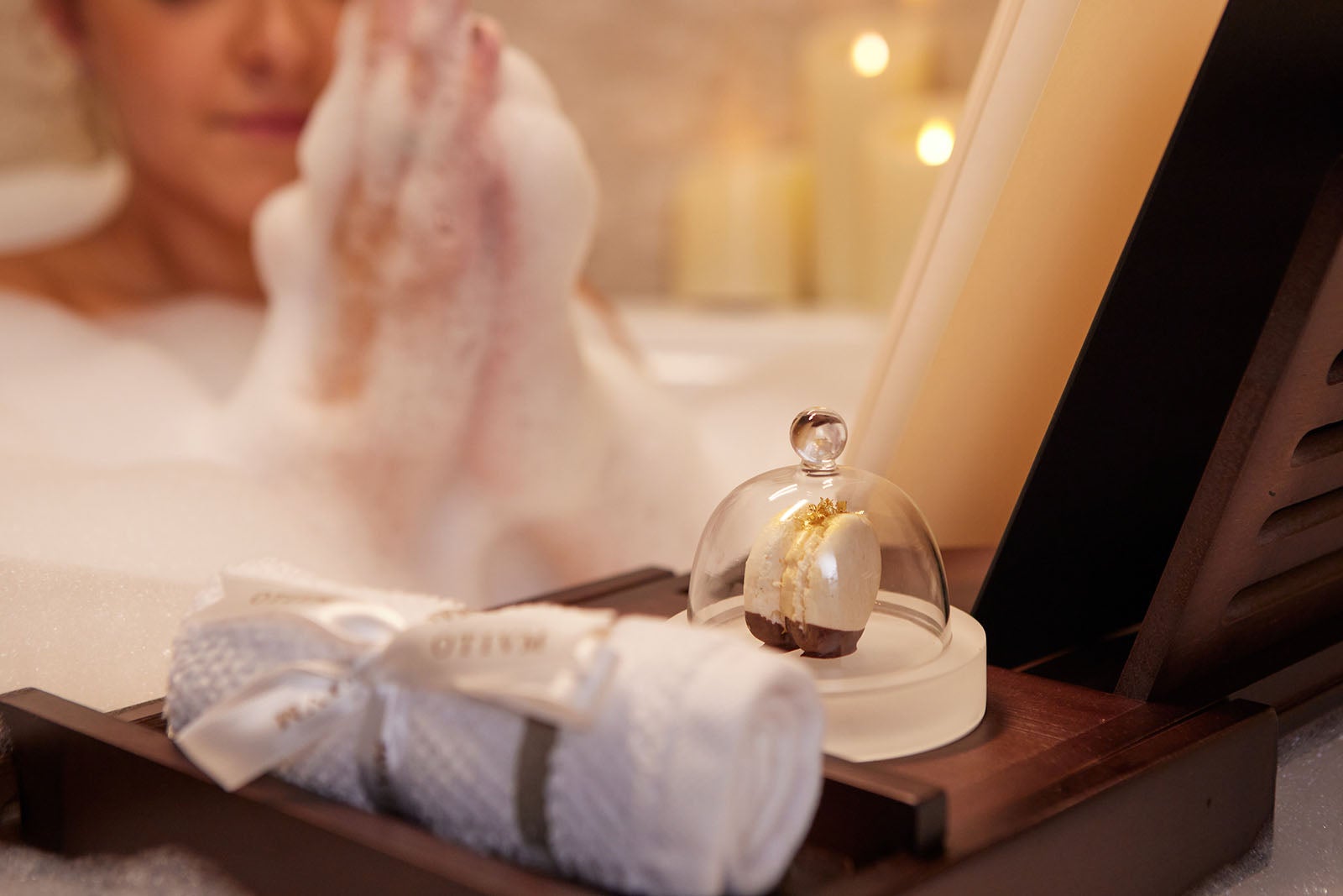 Woman in bubble bath with wooden tray holding a book, macaron under glass and rolled-up wash cloth