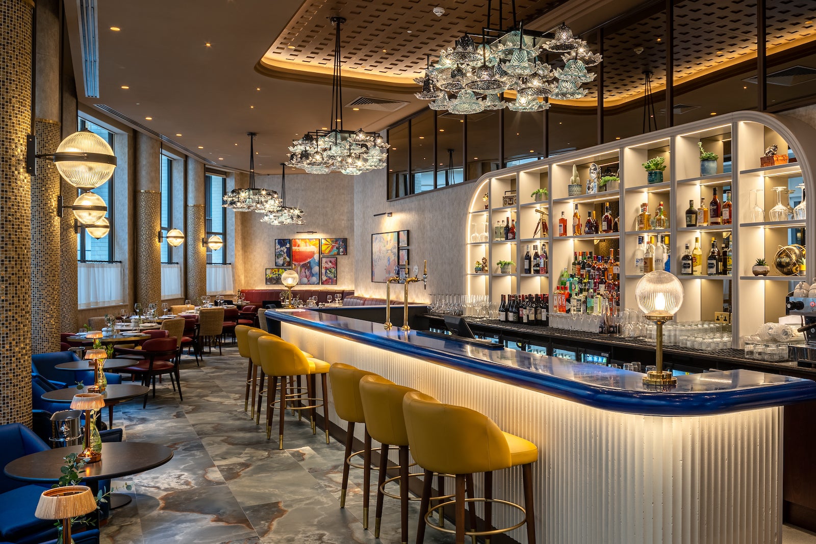 eclectic bar with blue counters, yellow bar height stools, tables against a wall and bottles of liquor on curved shelf