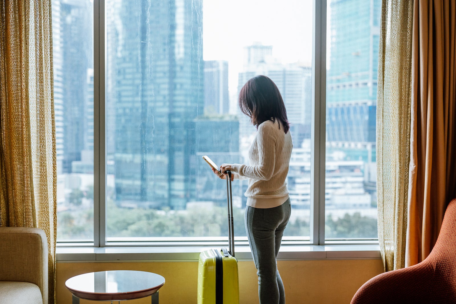 woman on phone with suitcase standing in front of window with city skyline in background