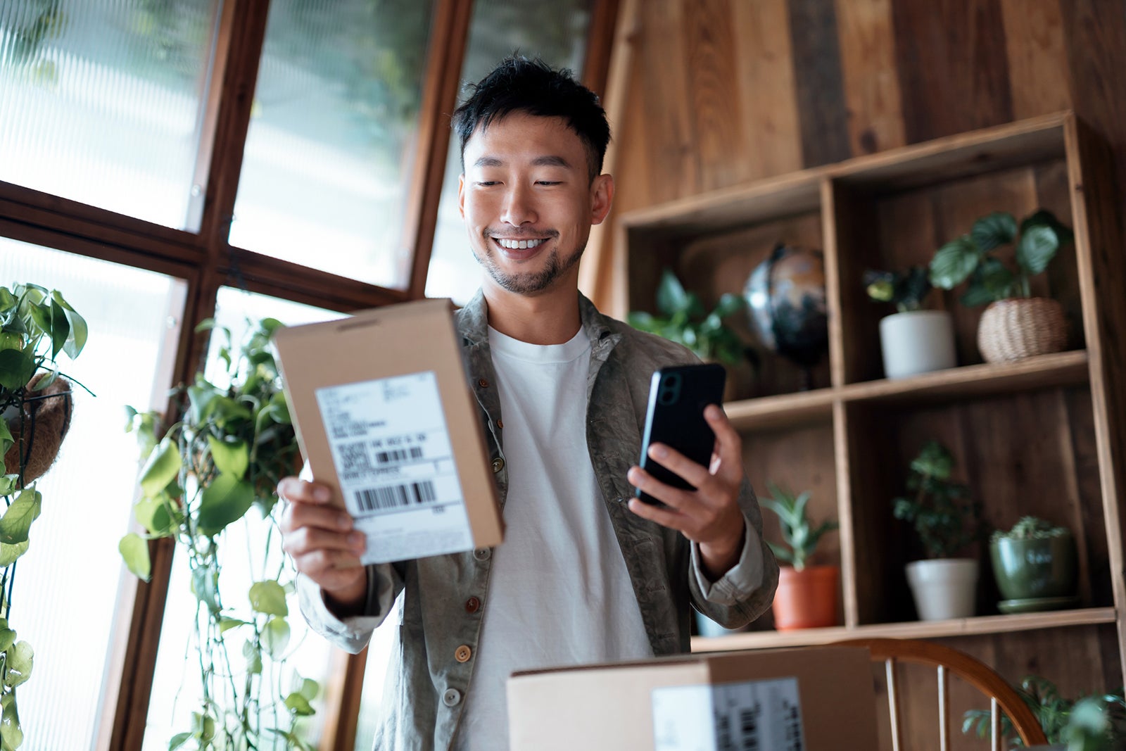 Smiling young Asian man checking electronic banking on his smartphone as he received delivered packages from online purchases at home. Online shopping. Online banking. Shopping and paying safely online