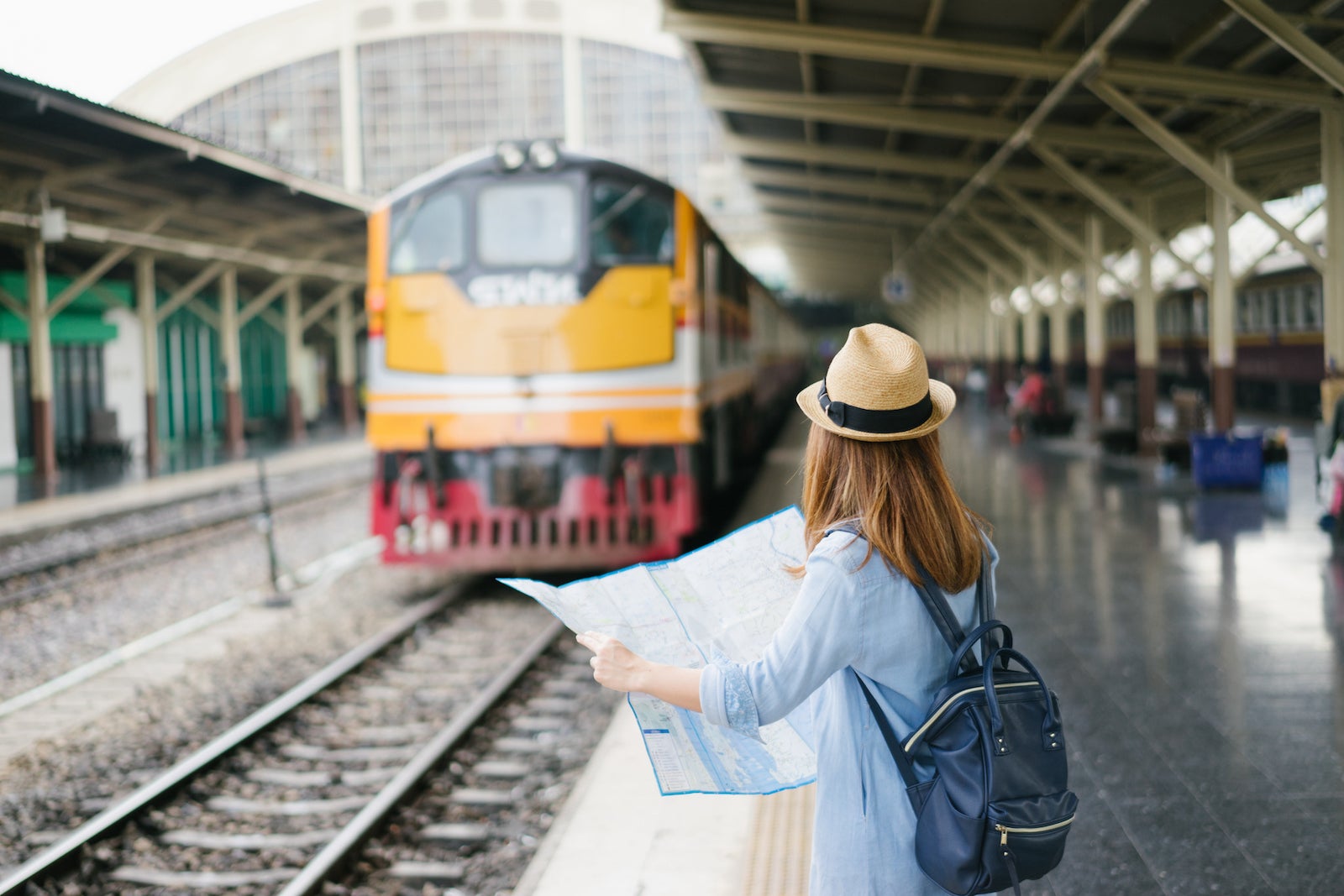 Young woman traveler with sky blue backpack and hat looking the map with train background at train station Bangkok. Traveling in Bangkok Thailand. Travel concept