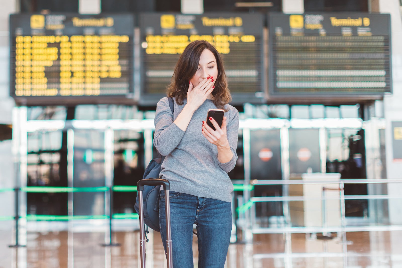 Image of excited, shocked young beautiful woman, standing in airport with smartphone in her hands - missed or cancelled flight concept - flight information board in background