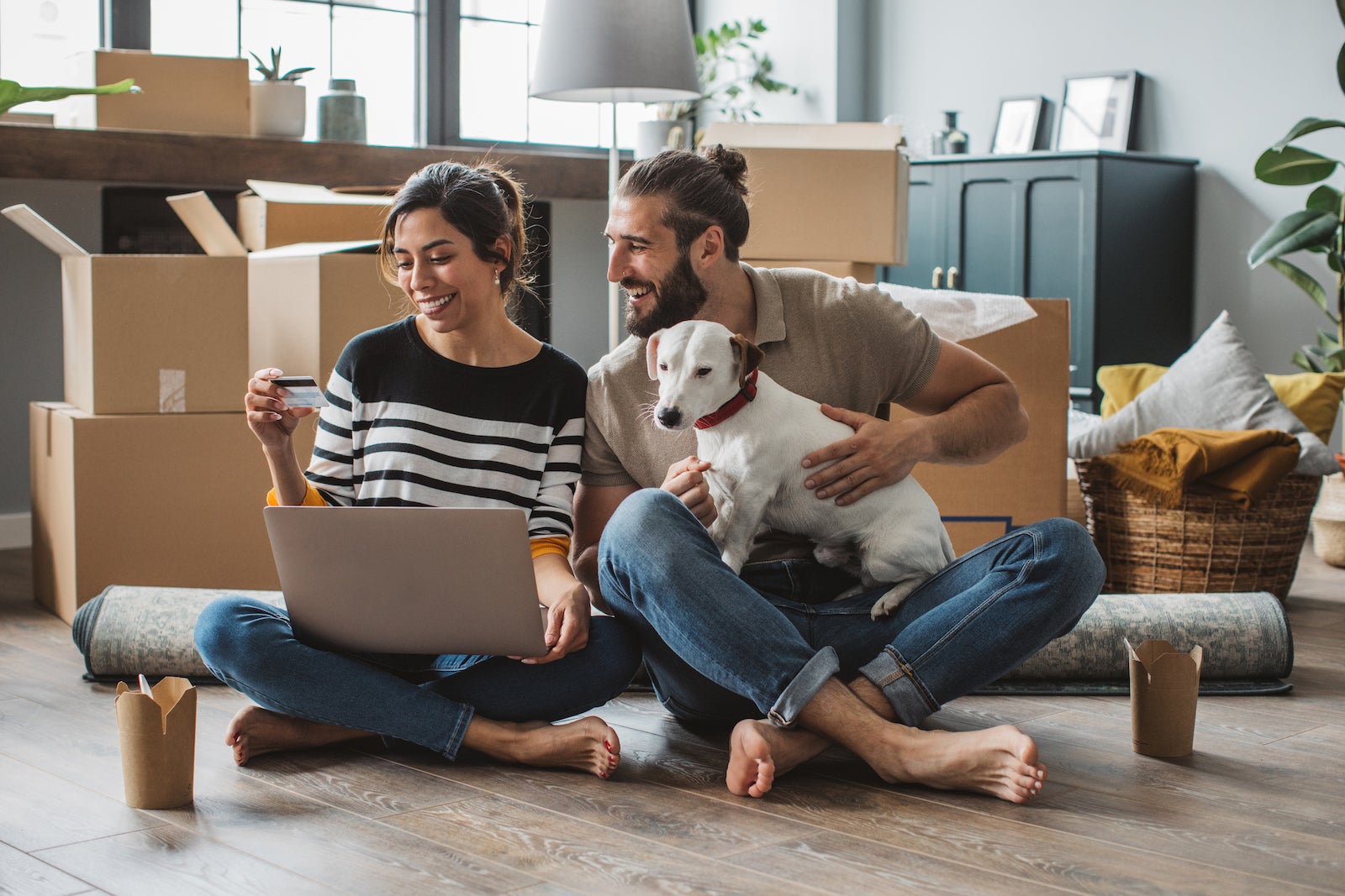 A young couple sits with a dog and moving boxes while shopping online with a laptop