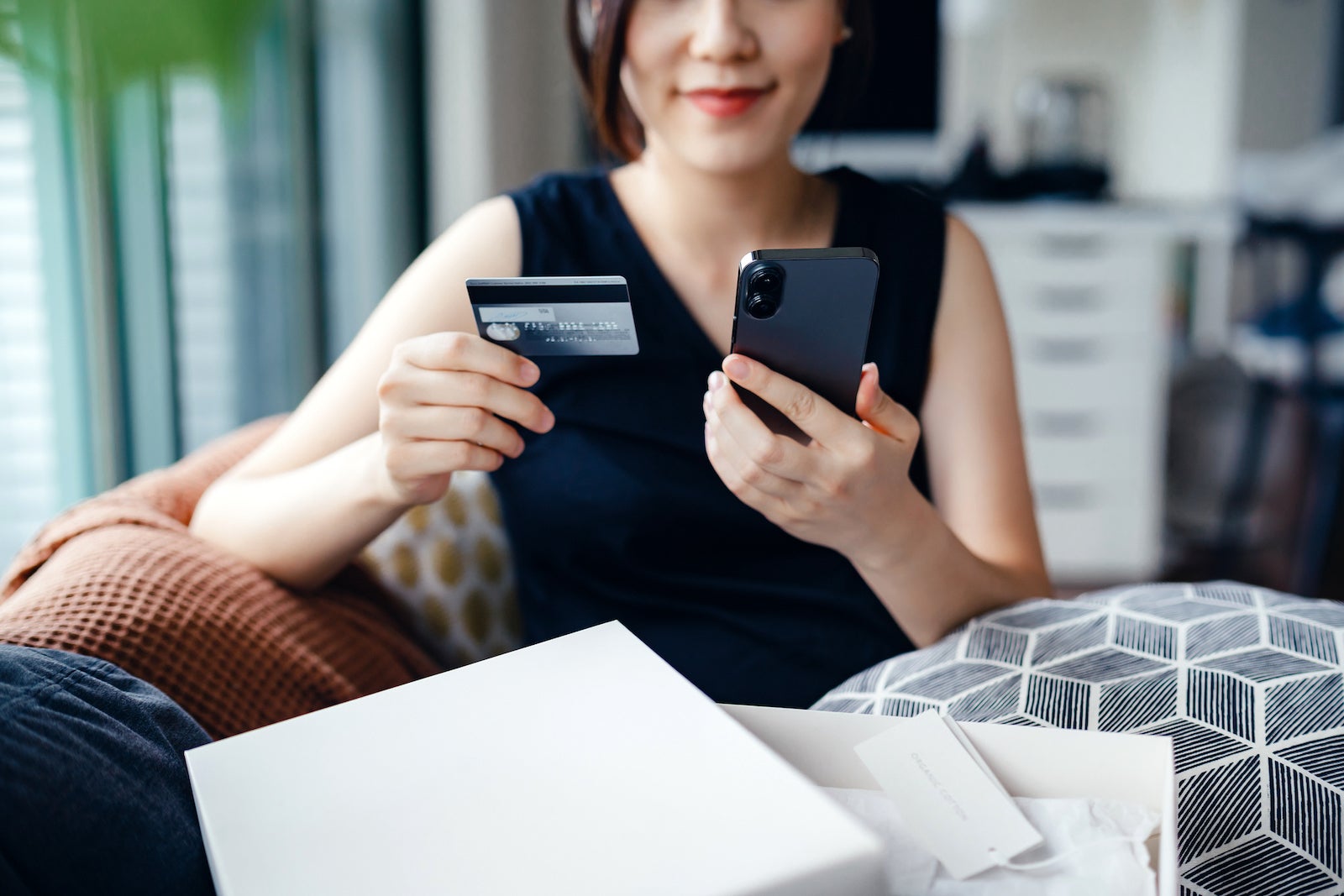 Beautiful smiling young Asian woman shopping online with smartphone and making payment online with credit card at home. With home delivery shopping parcels in front of her. Lifestyle and e-commerce concept. Convenience and easy online shopping experience