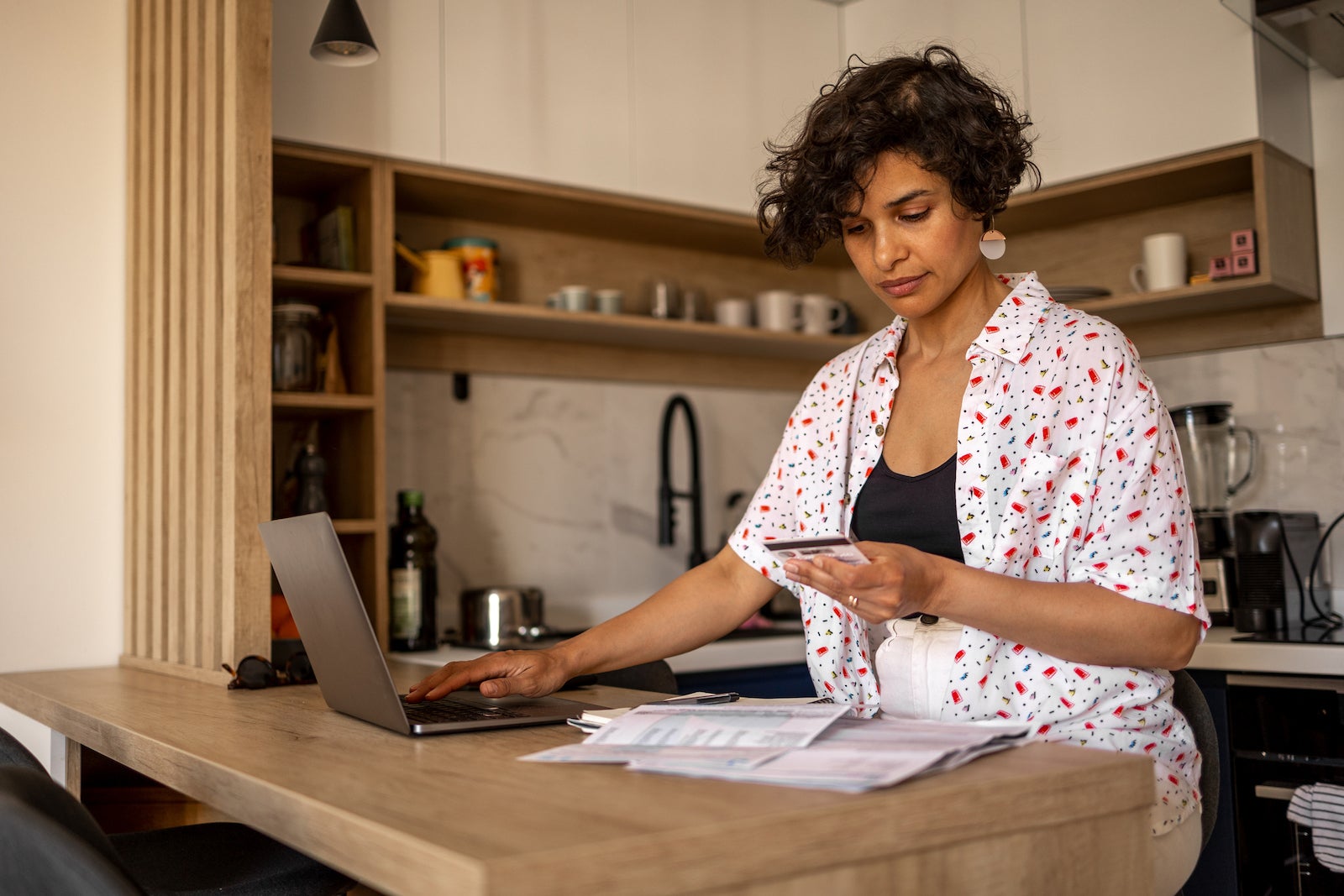 a woman looks at her credit card bills while using a laptop in her kitchen