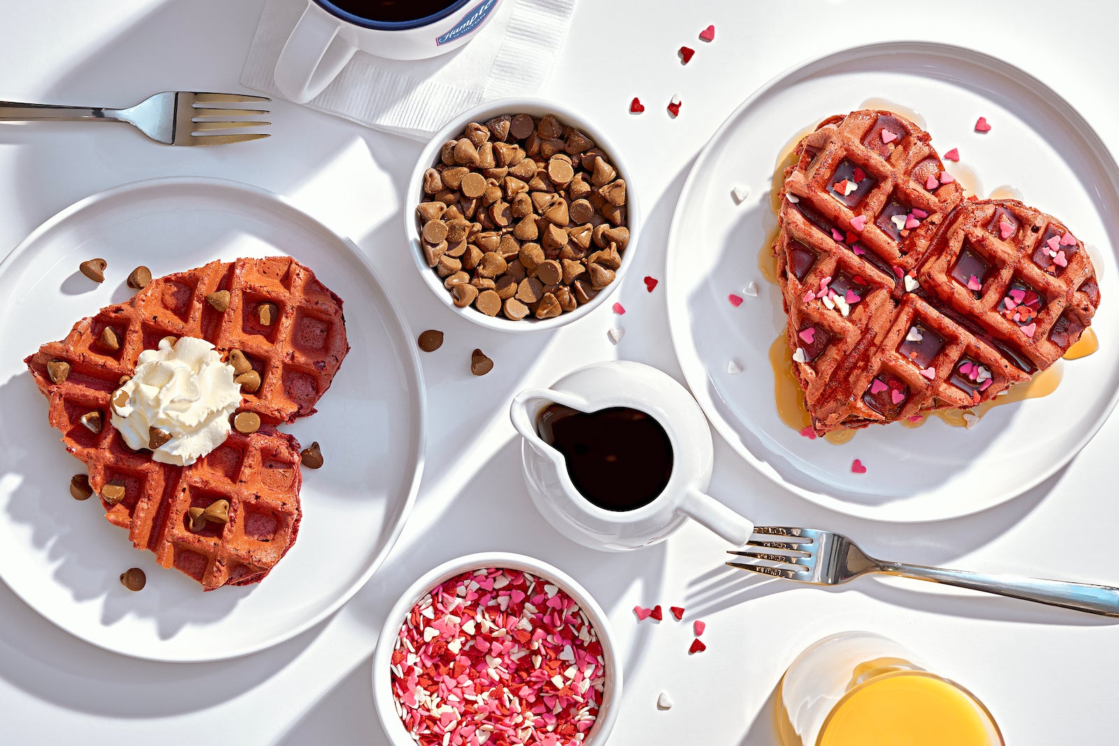 Heart-shaped pink waffles on white plates with bowls of chocolate chips, pink, red and white heart-shaped sprinkles, forks and coffee