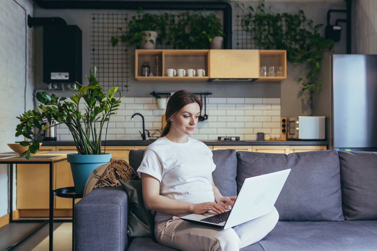 Maternity leave. Pregnant beautiful woman sitting on sofa at home with laptop in home clothes, ordering baby goods online, choosing, buying clothes and toys for baby.