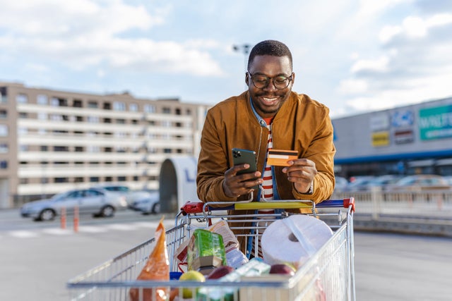 Portrait of a happy young African-American man pulling a shopping cart and using smart phone and credit card