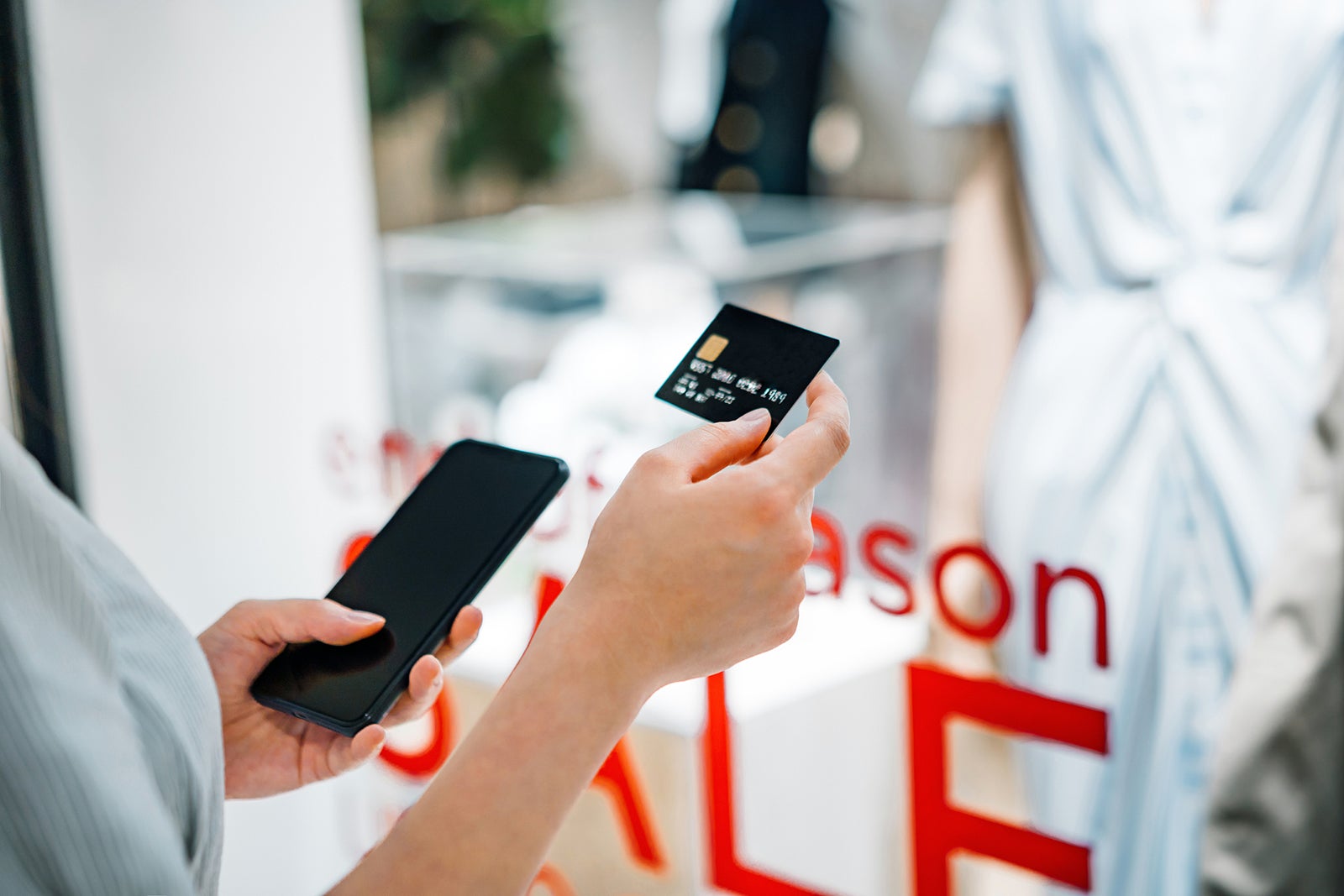 Cropped shot of young Asian woman holding a credit card and smartphone shopping in a department store, standing in front of a clothing store. Credit card and shopping concept