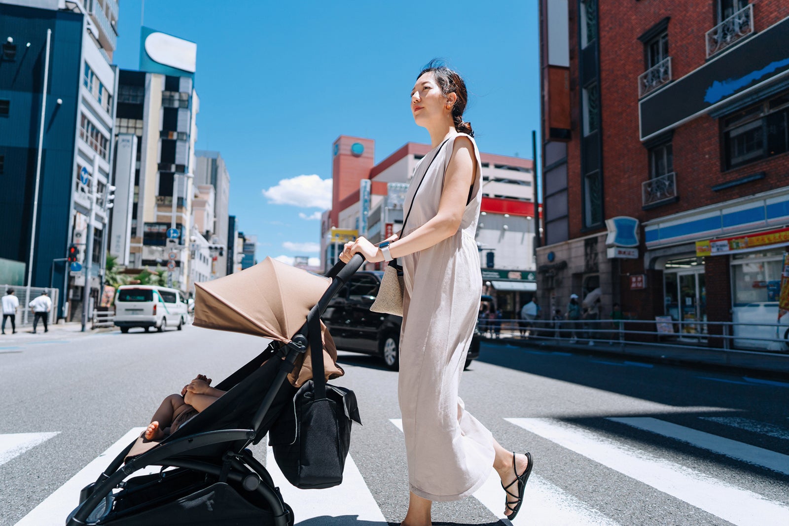 Young Asian mother with baby stroller walking down the city street, crossing the road on a sunny day. Family lifestyle. Family with baby